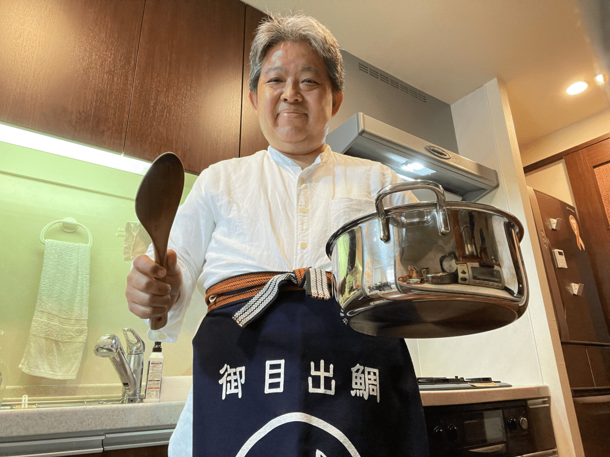 Yoshiki Ishiguro holding a pot and wooden spoon, wearing an apron with Japanese characters.
