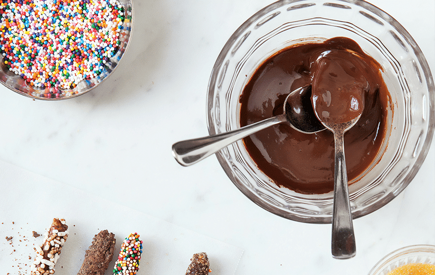A bowl of rich, glossy melted chocolate with sprinkles and chocolate sticks nearby.