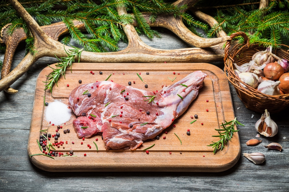 Venison steak on a wooden cutting board, flanked by fresh herbs, garlic, and rustic antlers.
